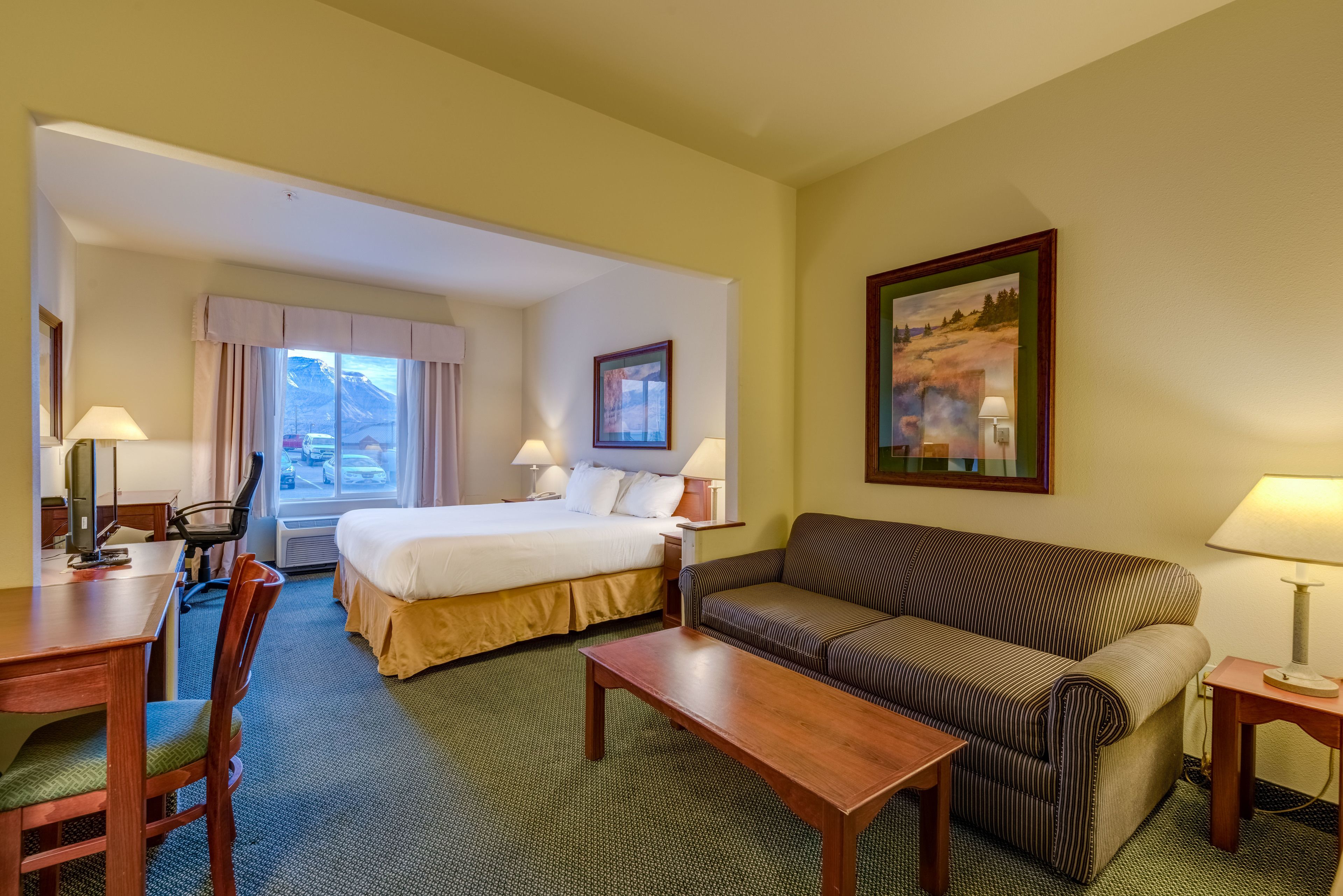 A hotel room with a bed, a sofa, a desk, and a chair. There are two lamps on either side of the bed and a painting on the wall.
