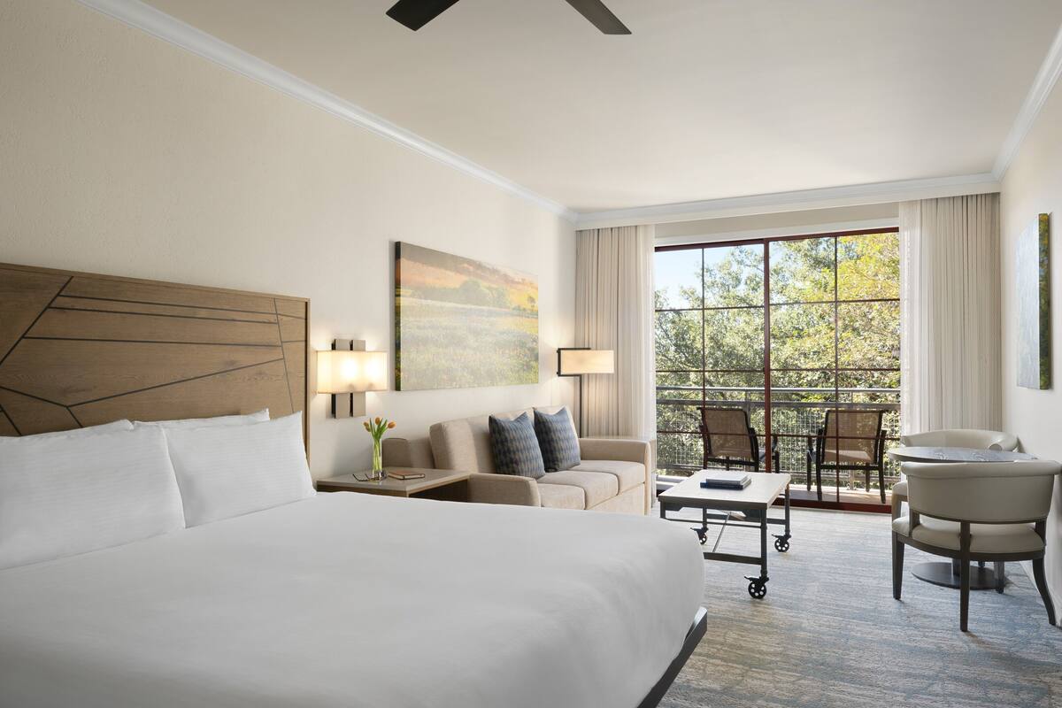 A hotel room with a large bed, a sofa, a desk, and a chair. There is a window with a view of trees and a balcony with a table and chairs.