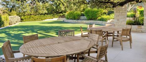 A round wooden table with chairs in a patio area with a pool and greenery in the background.