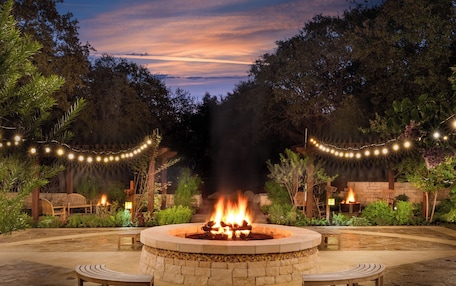 A circular stone fire pit with two wooden benches, surrounded by a stone patio and string lights.