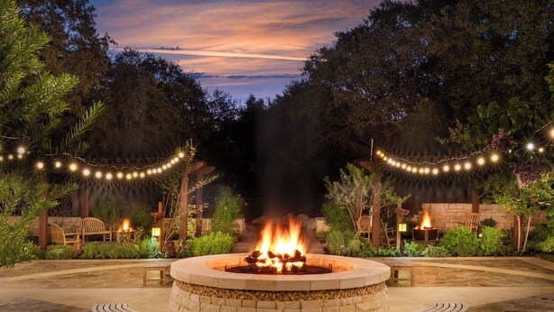 A circular stone fire pit with two wooden benches, surrounded by a stone patio and string lights. - Hyatt Vacation Club at Wild Oak Ranch, San Antonio (San Antonio)