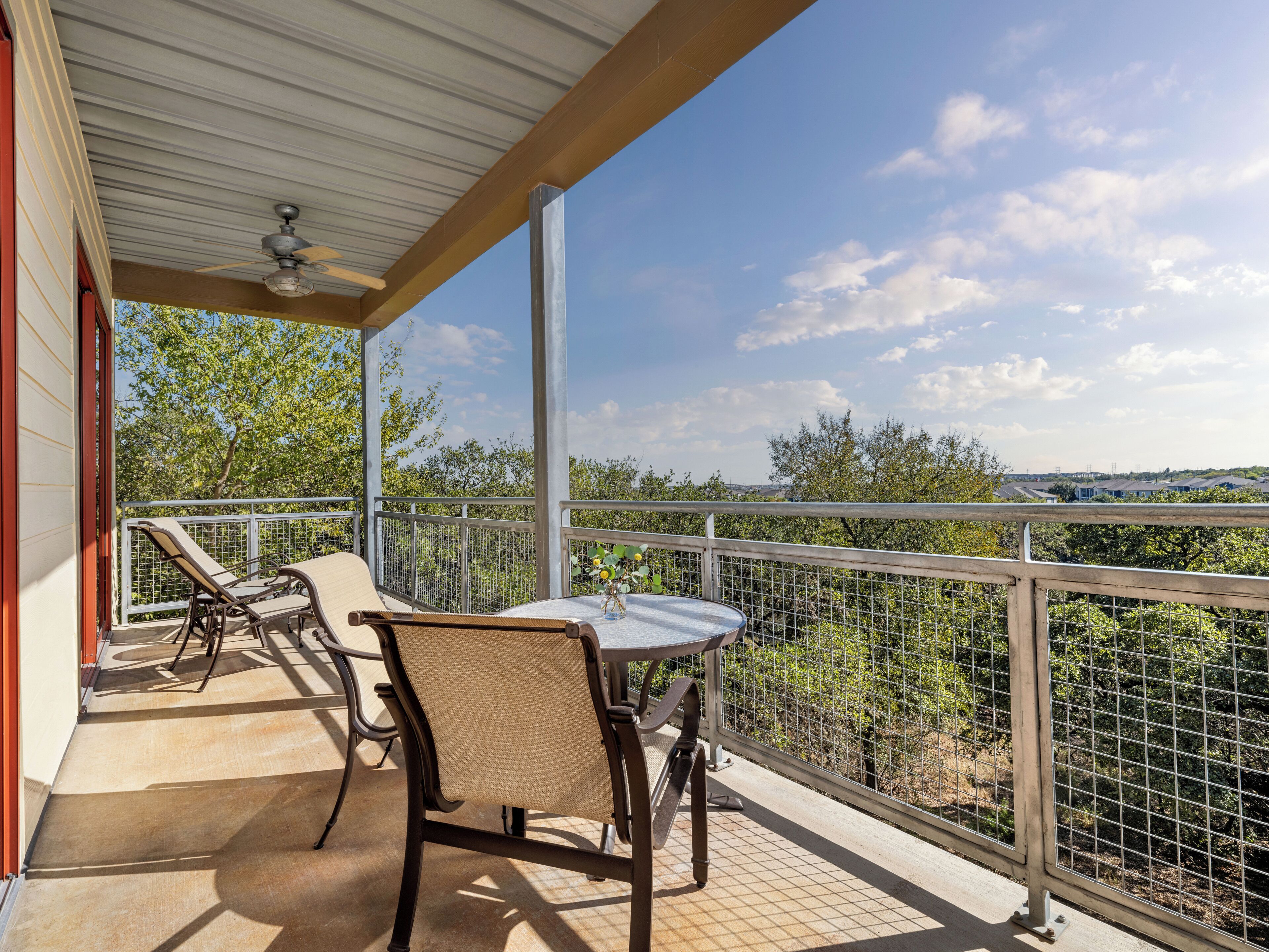 A covered balcony with a table and chairs, a ceiling fan, and a view of trees and buildings.