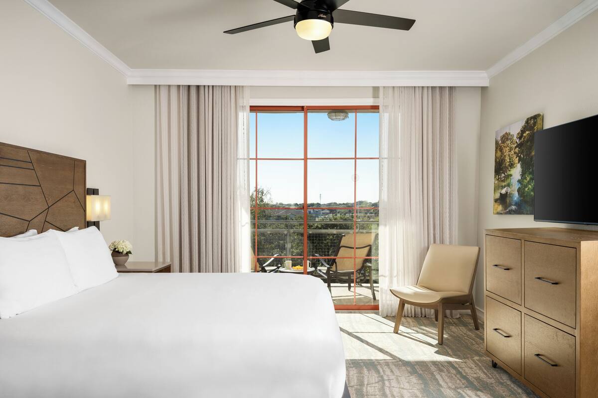 A hotel room with a large bed, a ceiling fan, a TV, a dining area with a table and chairs, and a view of the outdoors.