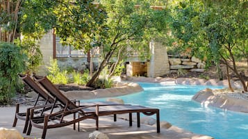 Zona de piscina con sillones de descanso, pared de piedra y piscina azul cristalina rodeada de vegetación.