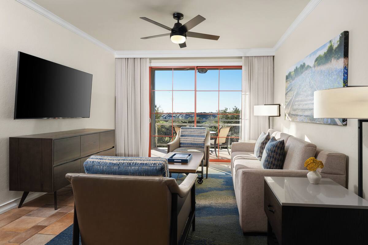 A hotel room with a flat-screen TV, a ceiling fan, a sofa, a chair, a small table, and a large window with a view of the outdoors.