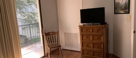 A room with a wooden dresser, a television on top, a chair, and a sliding glass door leading to a balcony.