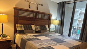 A bedroom with a wooden headboard, a checkered bedspread, two bedside lamps, a small plant, and a view of the balcony. - Oak Square Rentals (Gatlinburg)