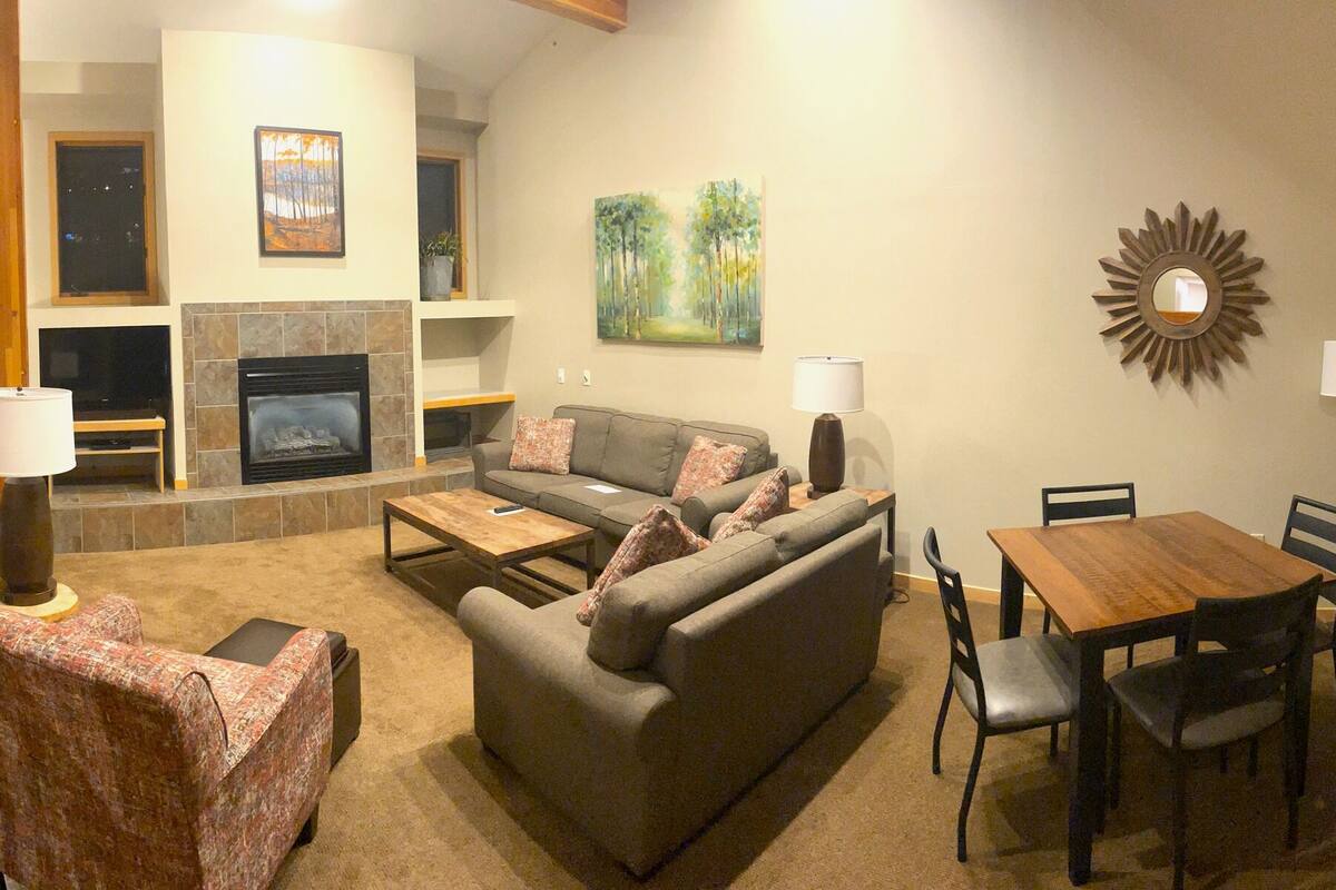 A spacious living area with a fireplace, a dining table, and a kitchen in the background.
