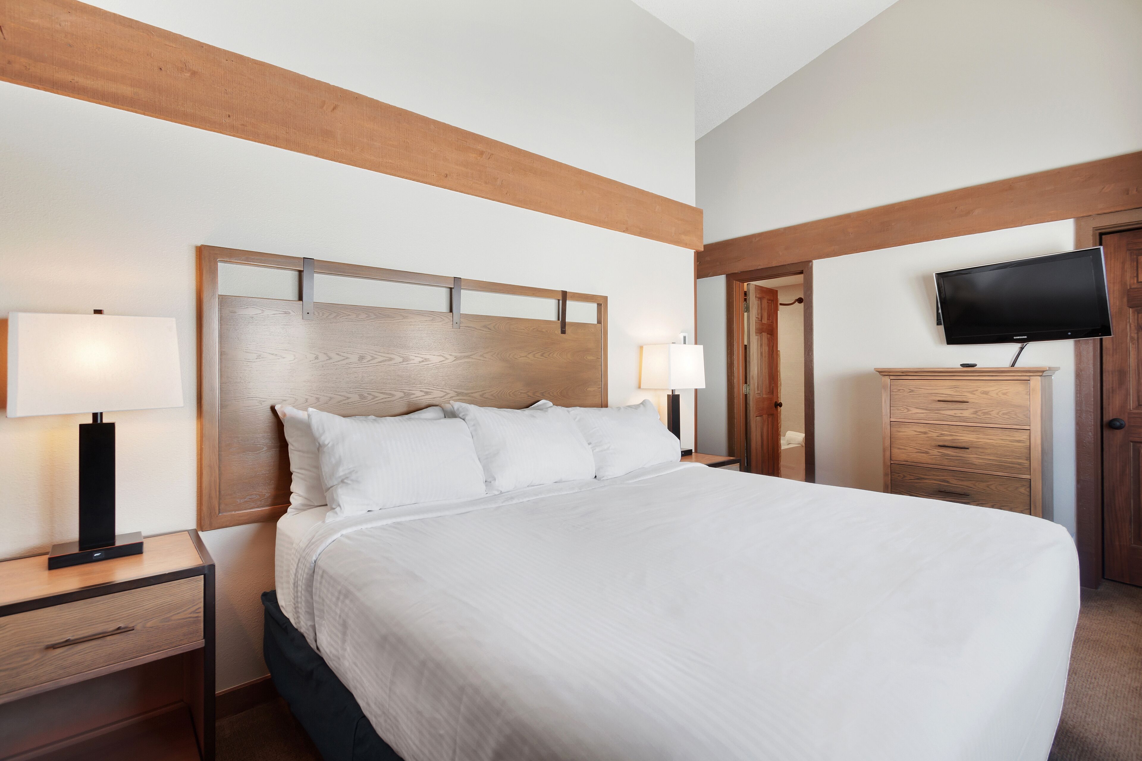 A hotel room with a large bed, wooden headboard, bedside tables with lamps, a TV on a wooden cabinet, and a door to another room.