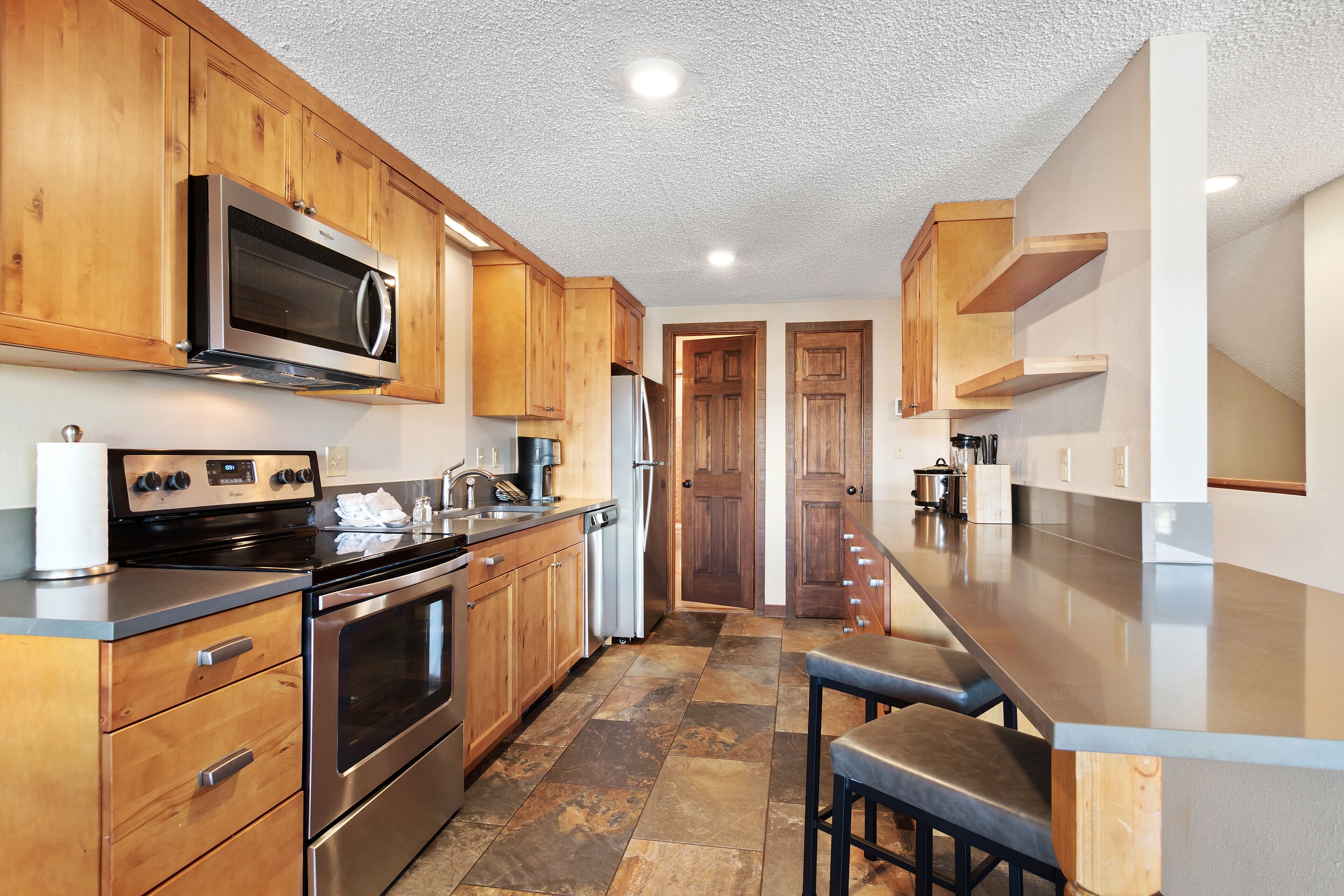 A modern kitchen with wooden cabinets, stainless steel appliances, and a stone tile floor.