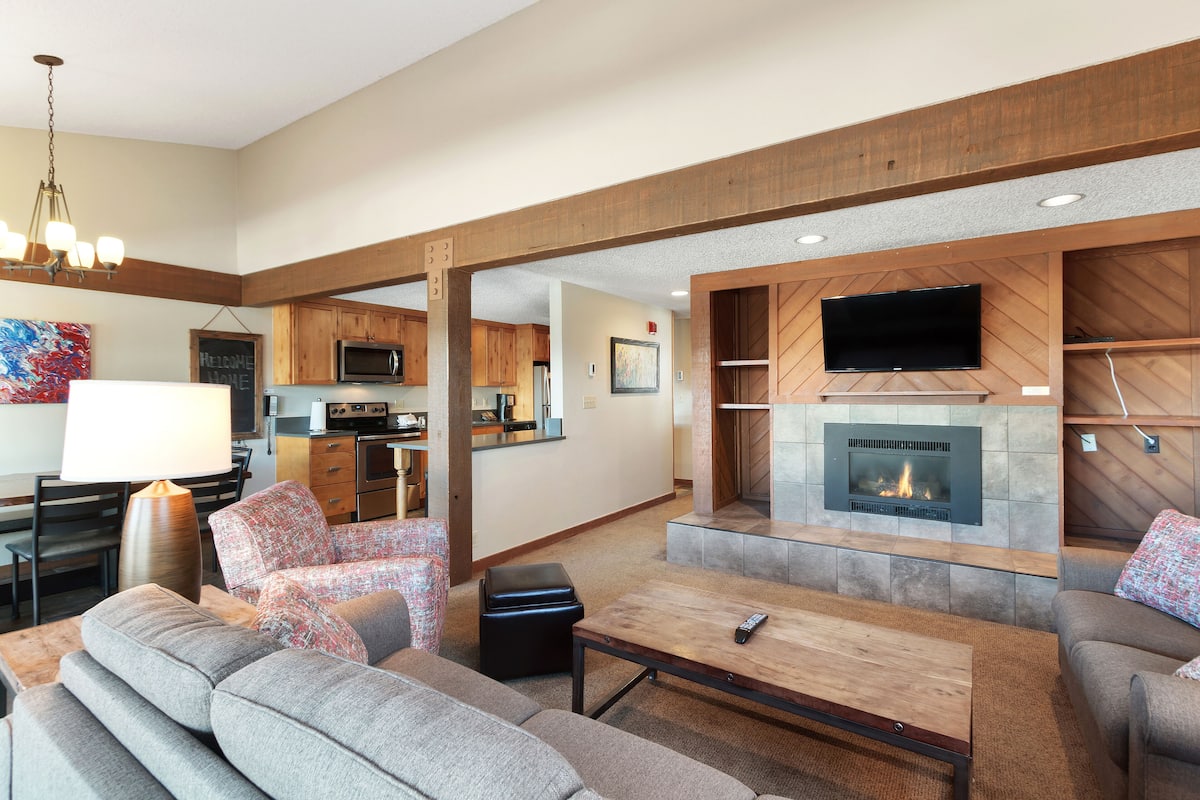 A spacious living room with a fireplace, a television mounted on the wall, and a kitchen area in the background.