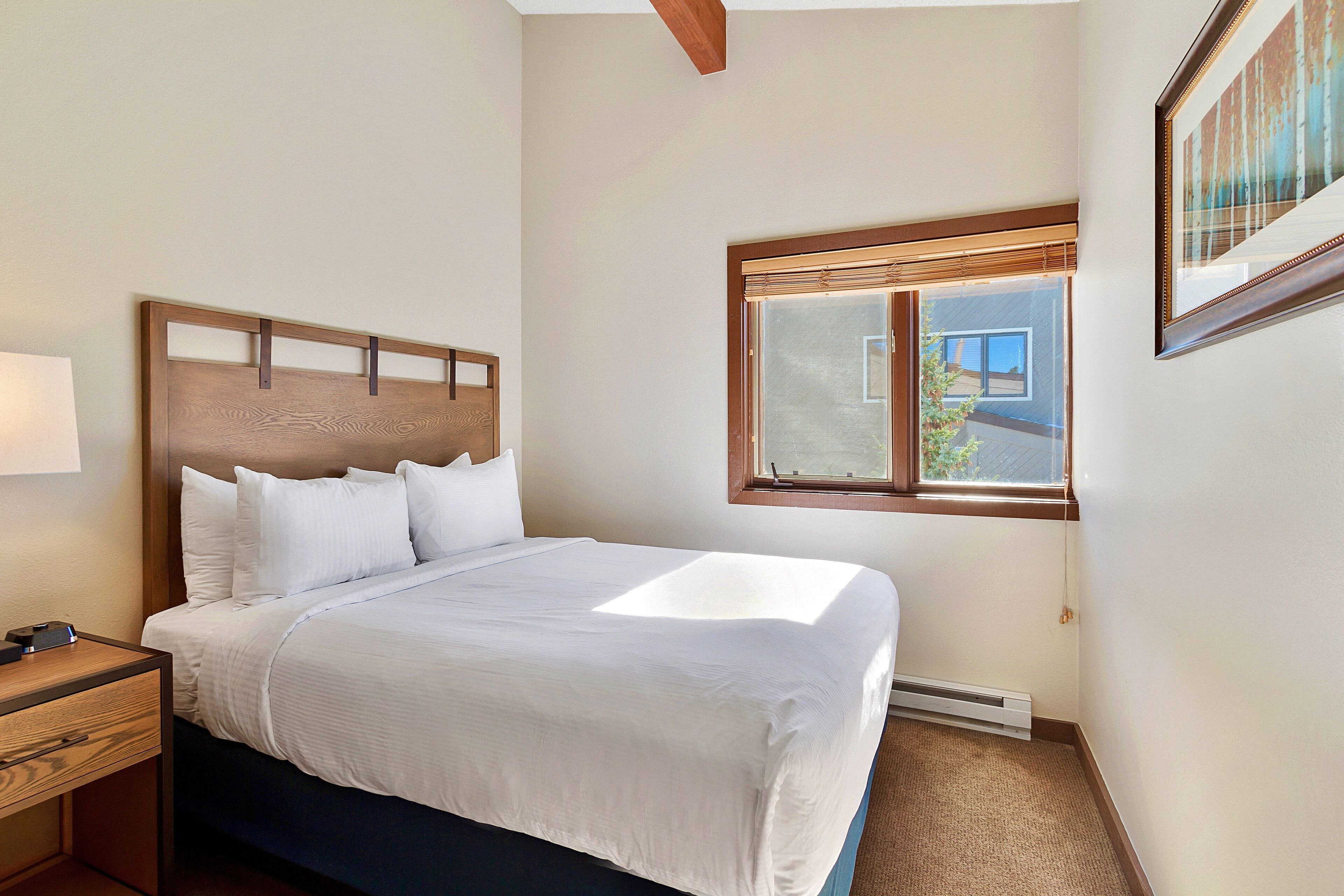 A neatly made bed with white linens, a wooden headboard, and a bedside table with a lamp. A window with blinds is visible, and a framed picture hangs on the wall.