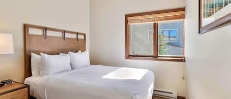 A neatly made bed with white linens, a wooden headboard, and a bedside table with a lamp. A window with blinds is visible, and a framed picture hangs on the wall.