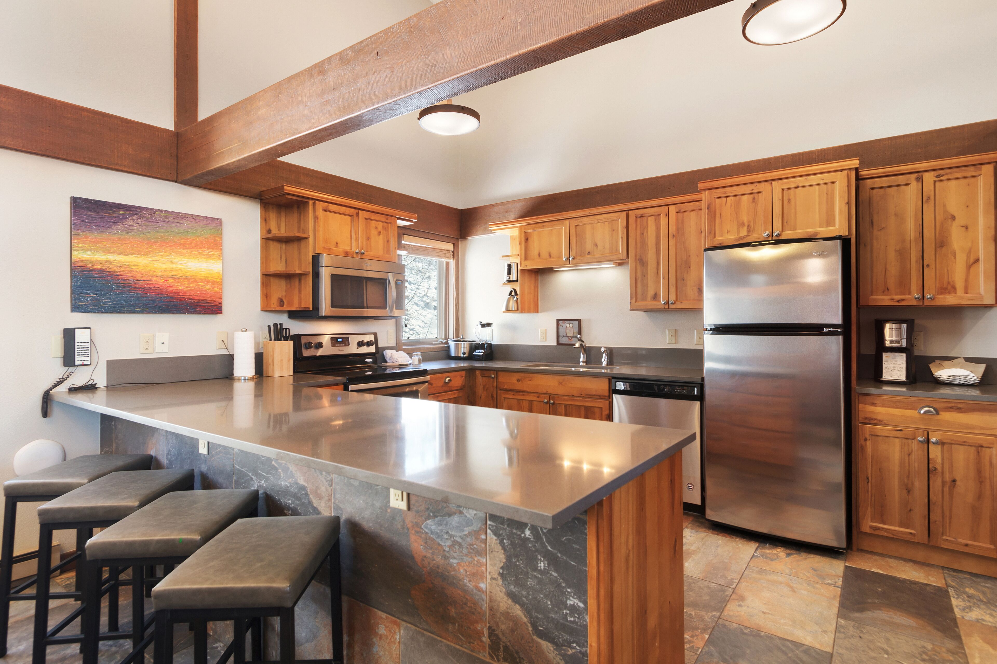 A modern kitchen with wooden cabinets, a large island with bar stools, stainless steel appliances, and a painting of a sunset on the wall.