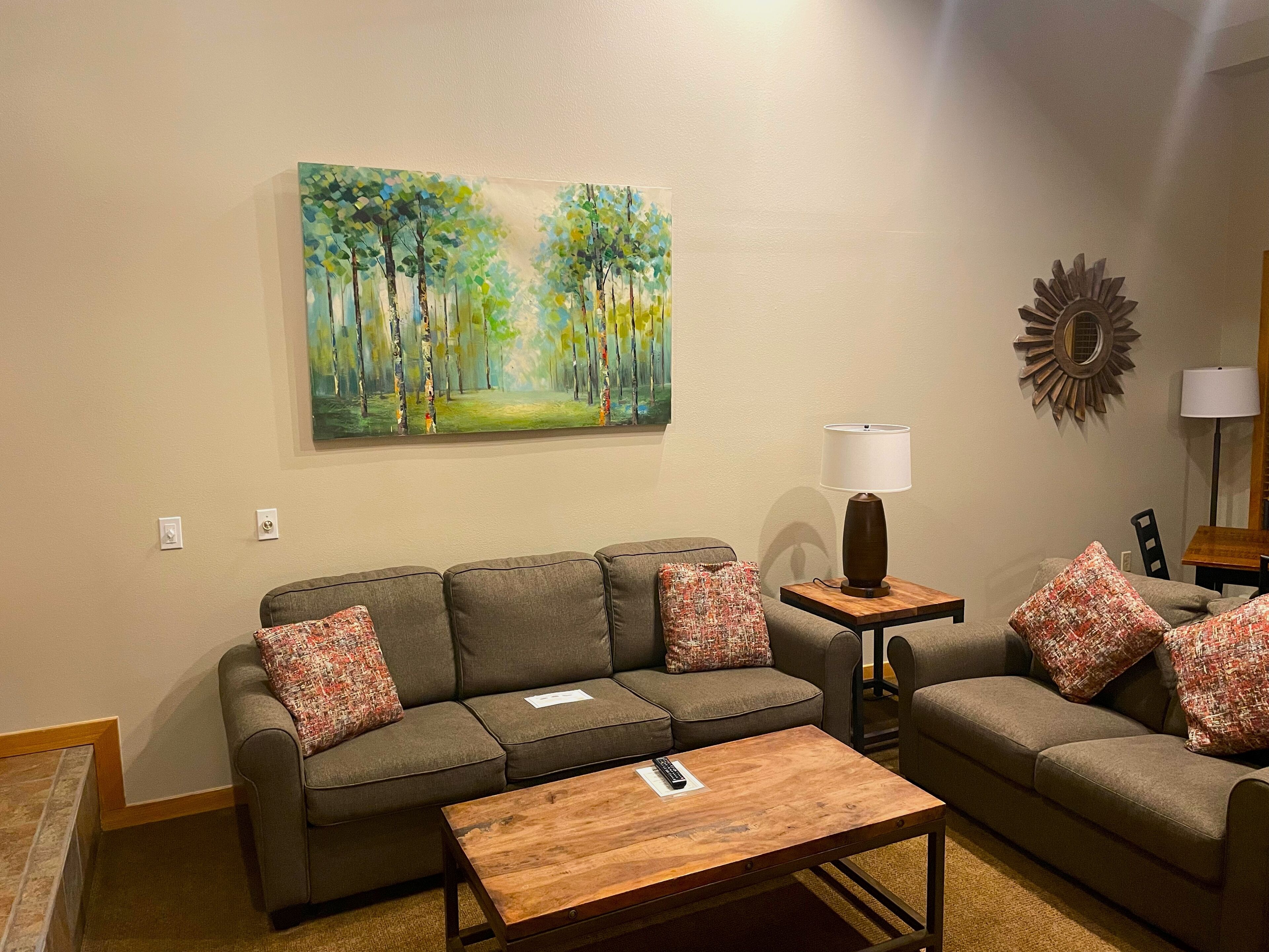 Un salon avec un canapé, une table basse, une lampe et un tableau représentant une forêt accroché au mur.