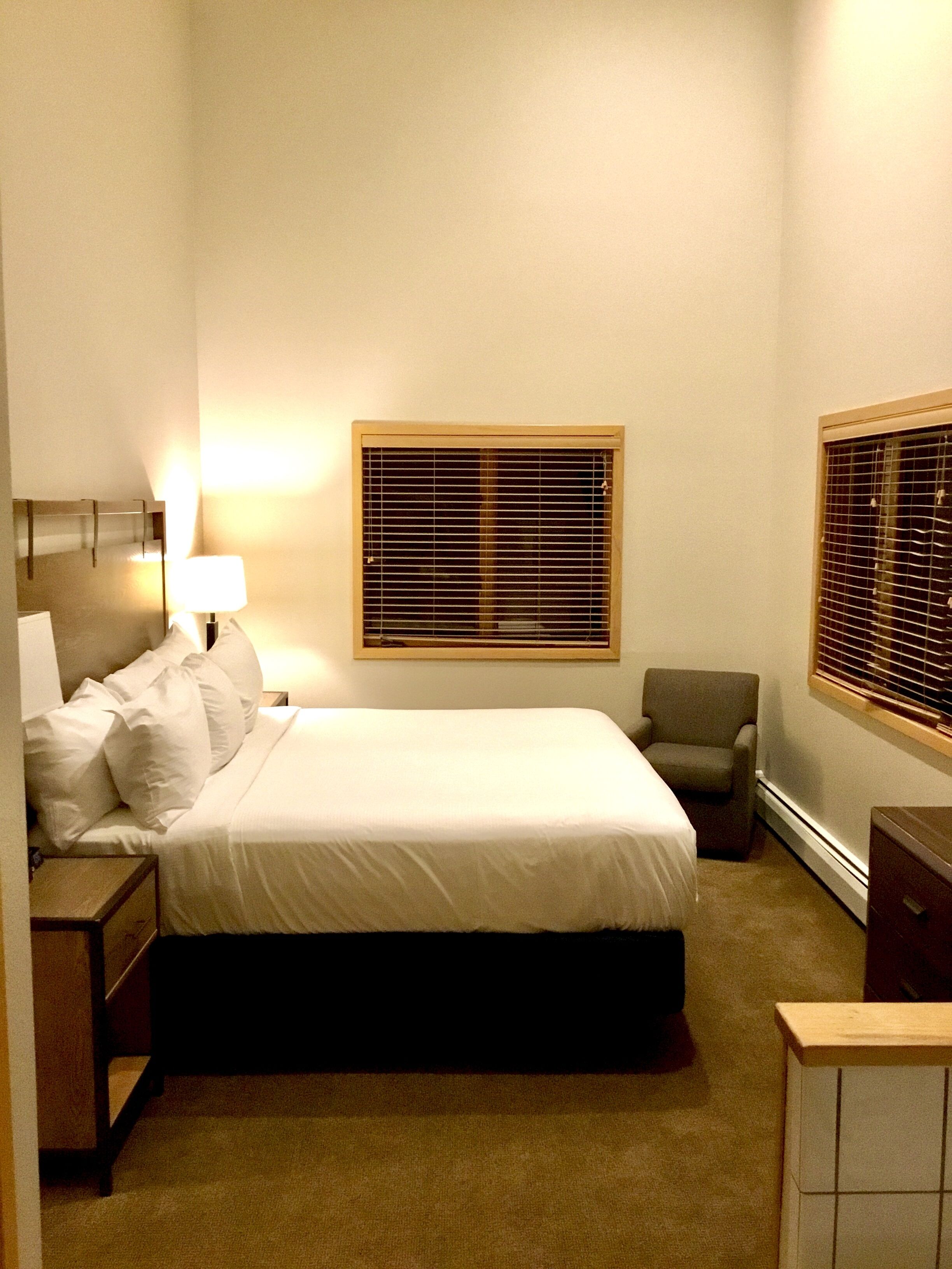 A hotel room with a bed, a chair, a nightstand, a lamp, and a window with blinds.