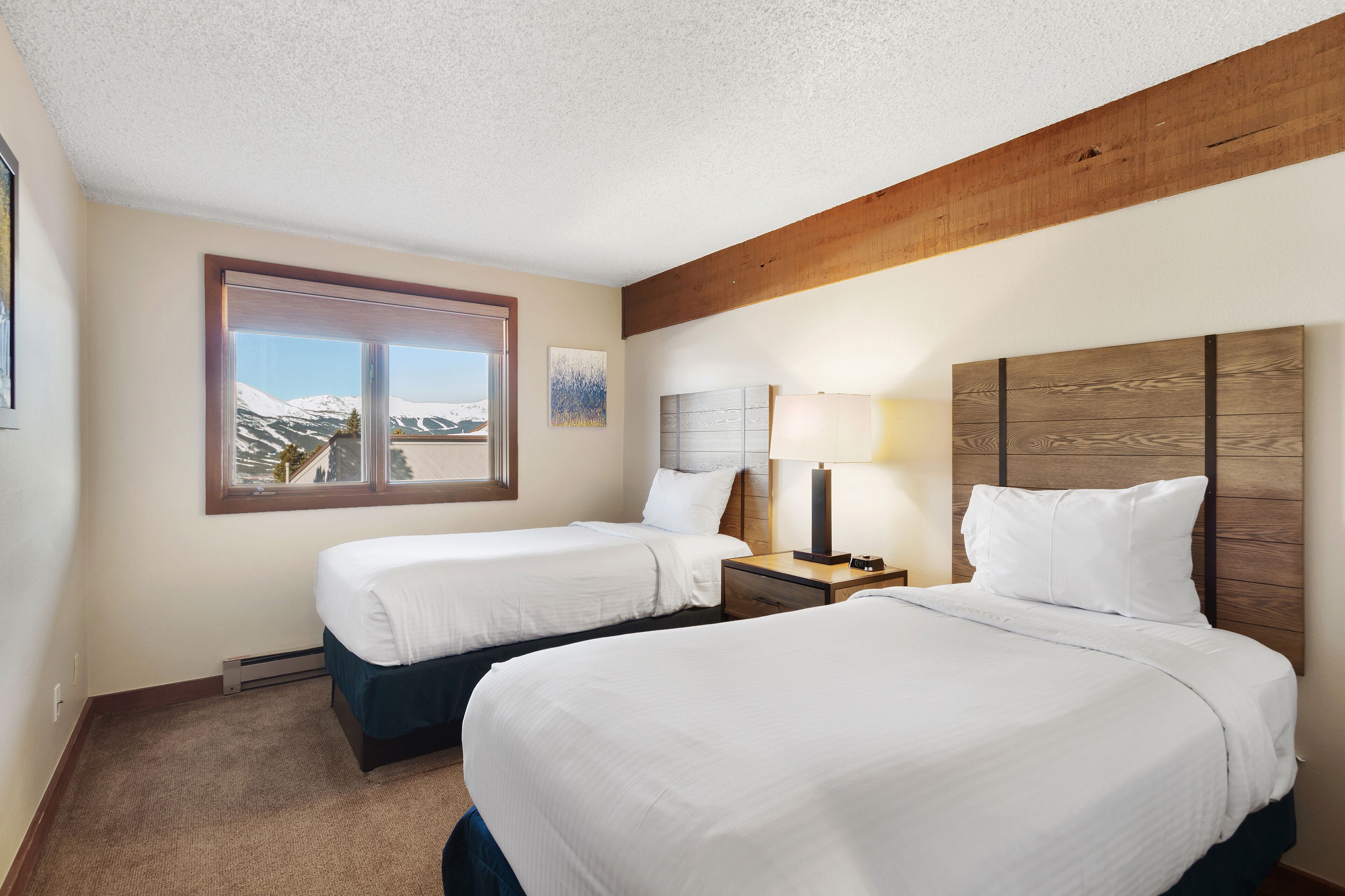 Une chambre d’hôtel avec deux lits, une fenêtre donnant sur les montagnes, une poutre en bois au plafond et une table de chevet avec une lampe.