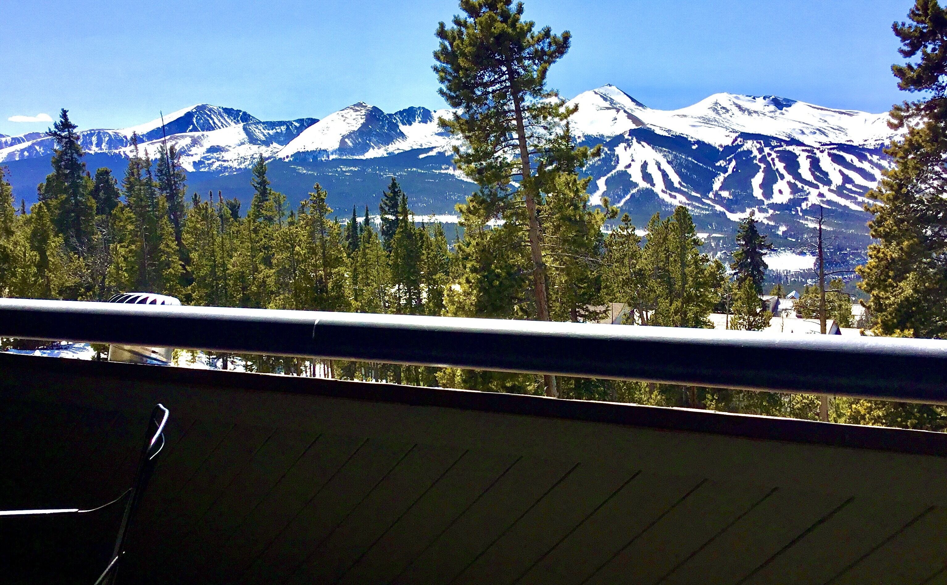 A mountain view with snow-capped peaks, a forest of pine trees, and a balcony railing.