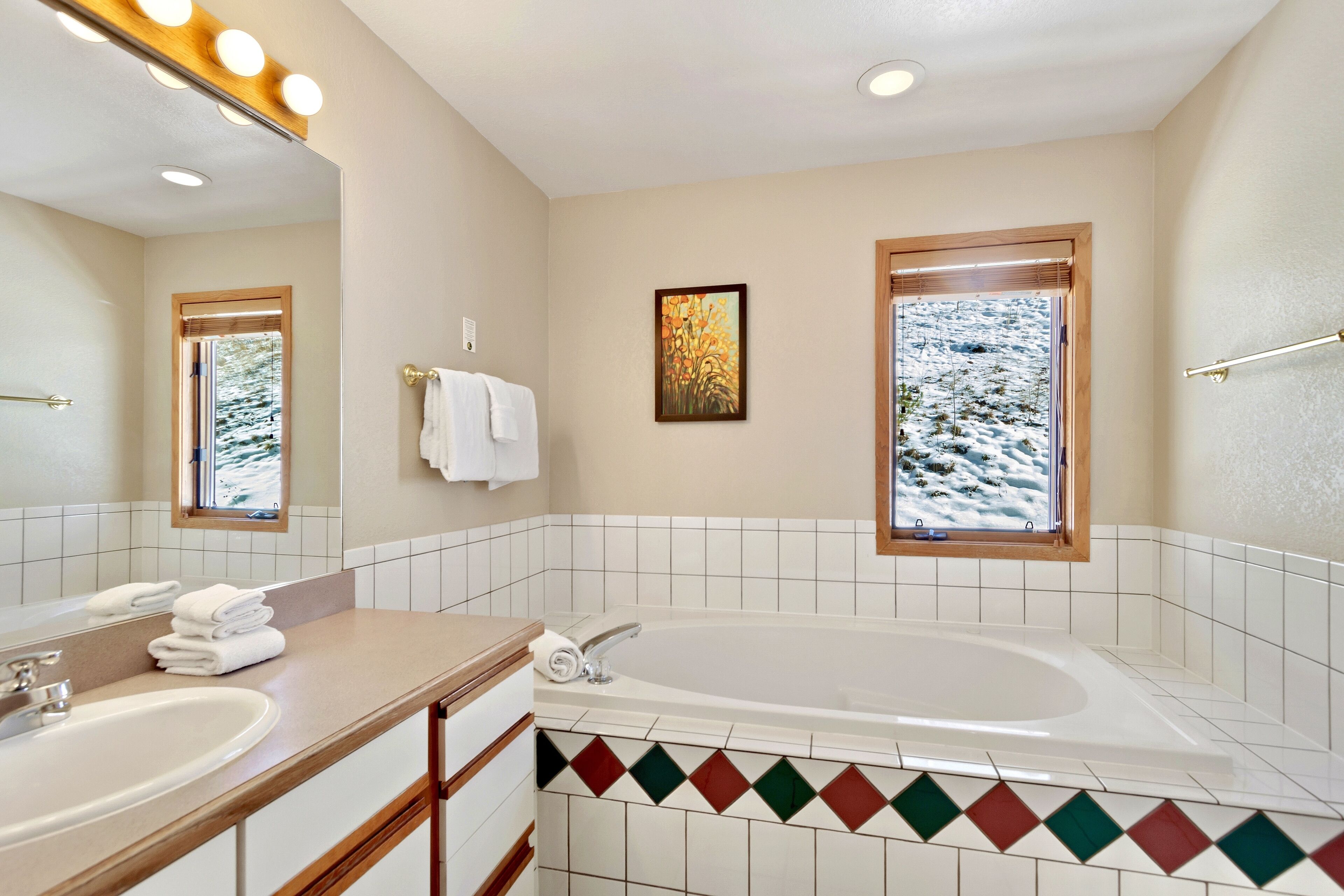 A bathroom with two sinks, a large bathtub, a mirror with lights, a window with a view of snow, and a painting on the wall.