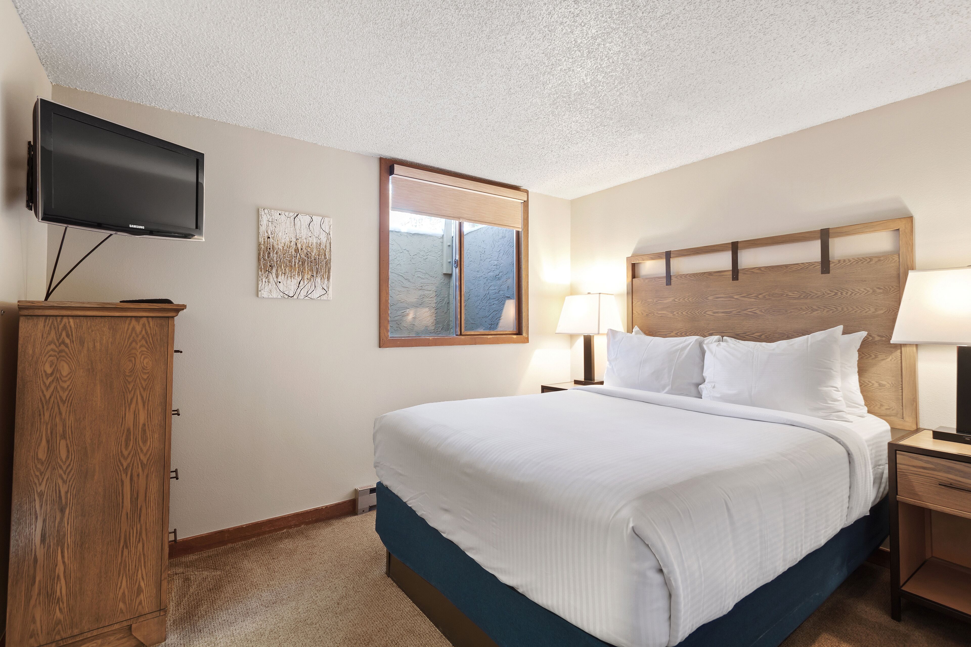 A hotel room with a large bed, a flat-screen TV mounted on the wall, a wooden headboard, and a nightstand with a lamp.