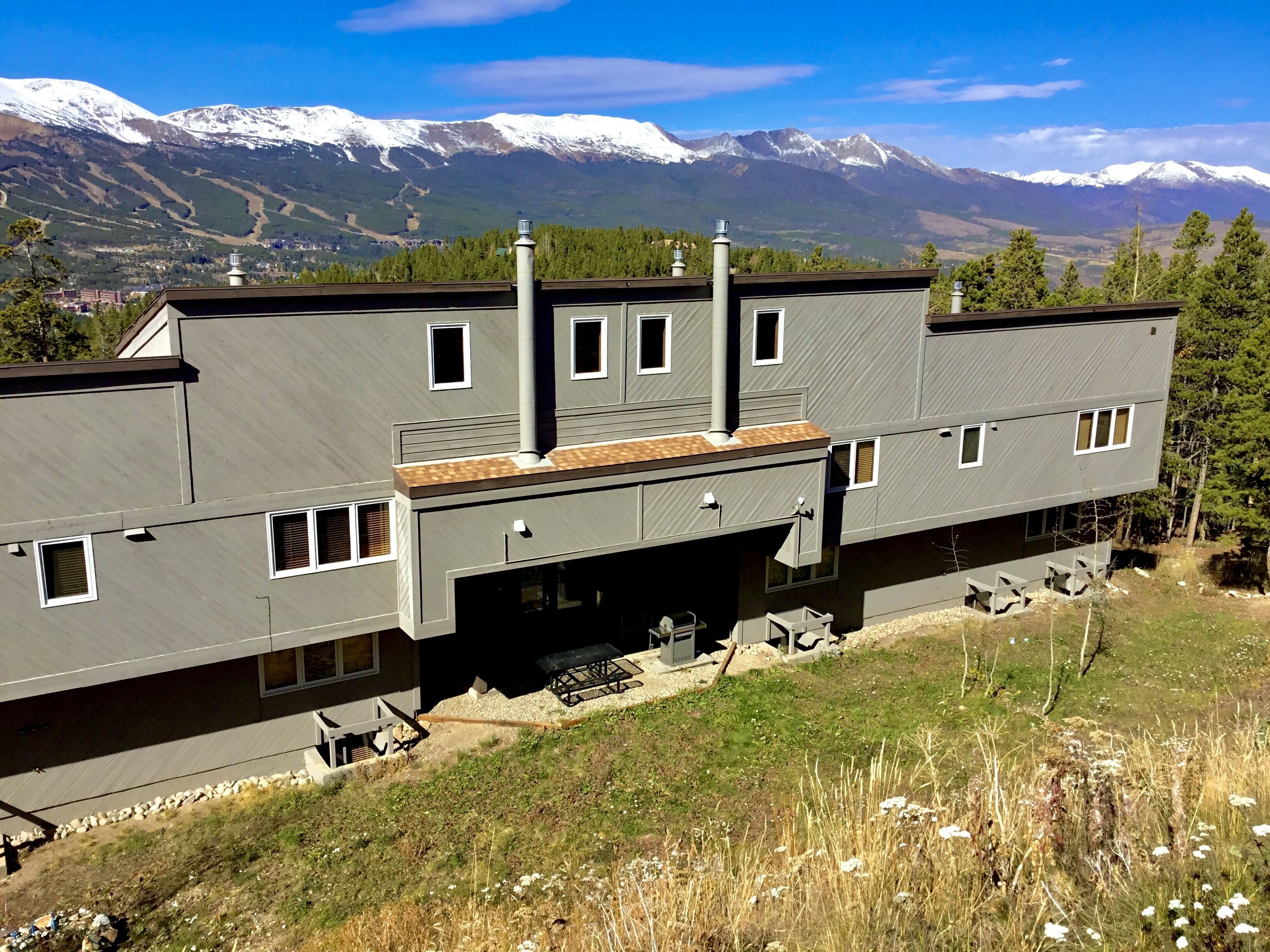 A modern two-story building with a flat roof, multiple windows, and a chimney. It is situated on a grassy hill with a view of snow-capped mountains in the background.