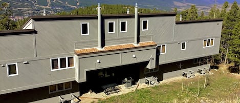 A modern two-story building with a flat roof, multiple windows, and a chimney. It is situated on a grassy hill with a view of snow-capped mountains in the background.