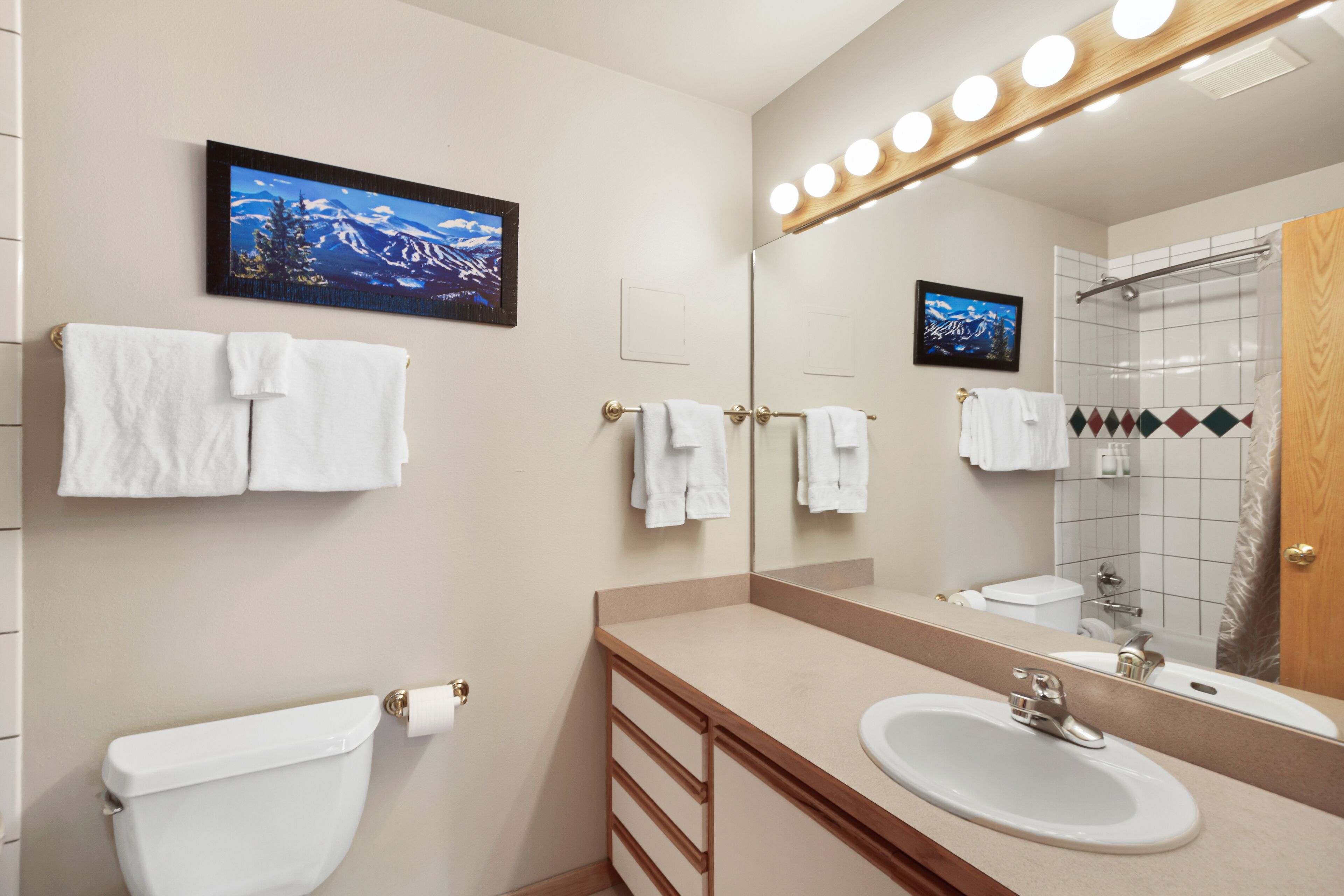 Une salle de bain avec des toilettes, un lavabo et un miroir. Plusieurs serviettes sont accrochées au mur, et il y a un espace douche avec une porte en verre.