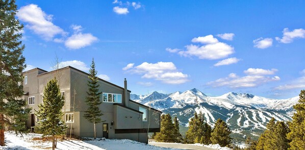 A modern two-story house with a ski slope view, surrounded by snow-covered mountains and evergreen trees.