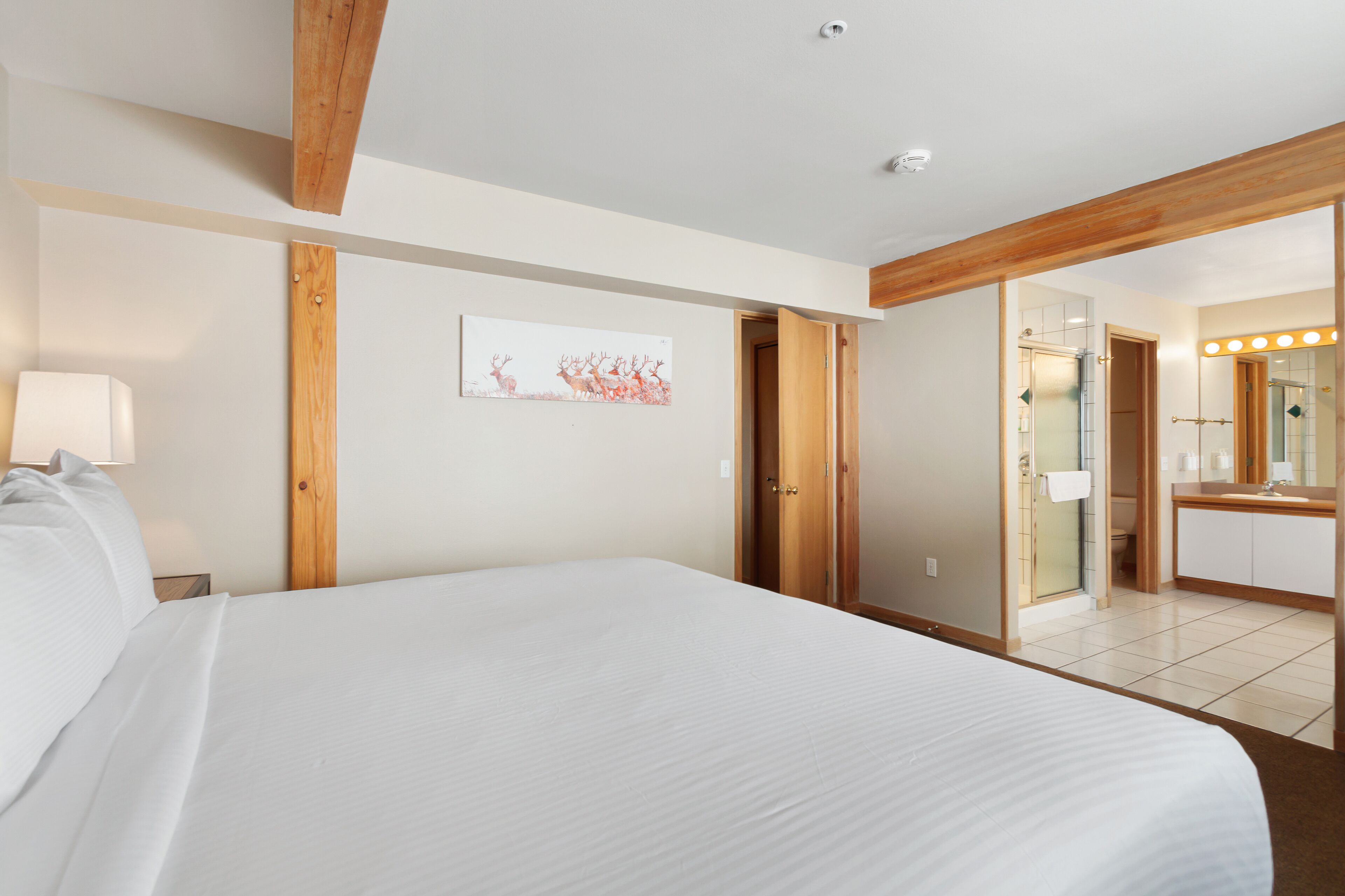 A hotel room with a large bed, a wooden ceiling, a bathroom with a mirror and lights, and a painting on the wall.