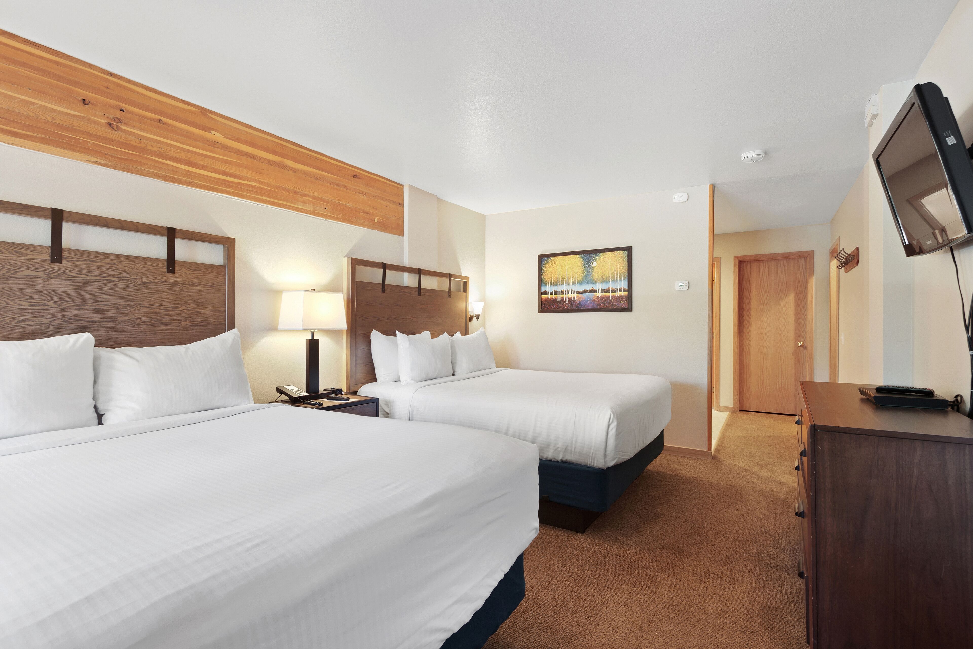 Habitación de hotel con dos camas, un cabecero de madera, un cuadro en la pared, un escritorio con televisor y una puerta que da a otra habitación.