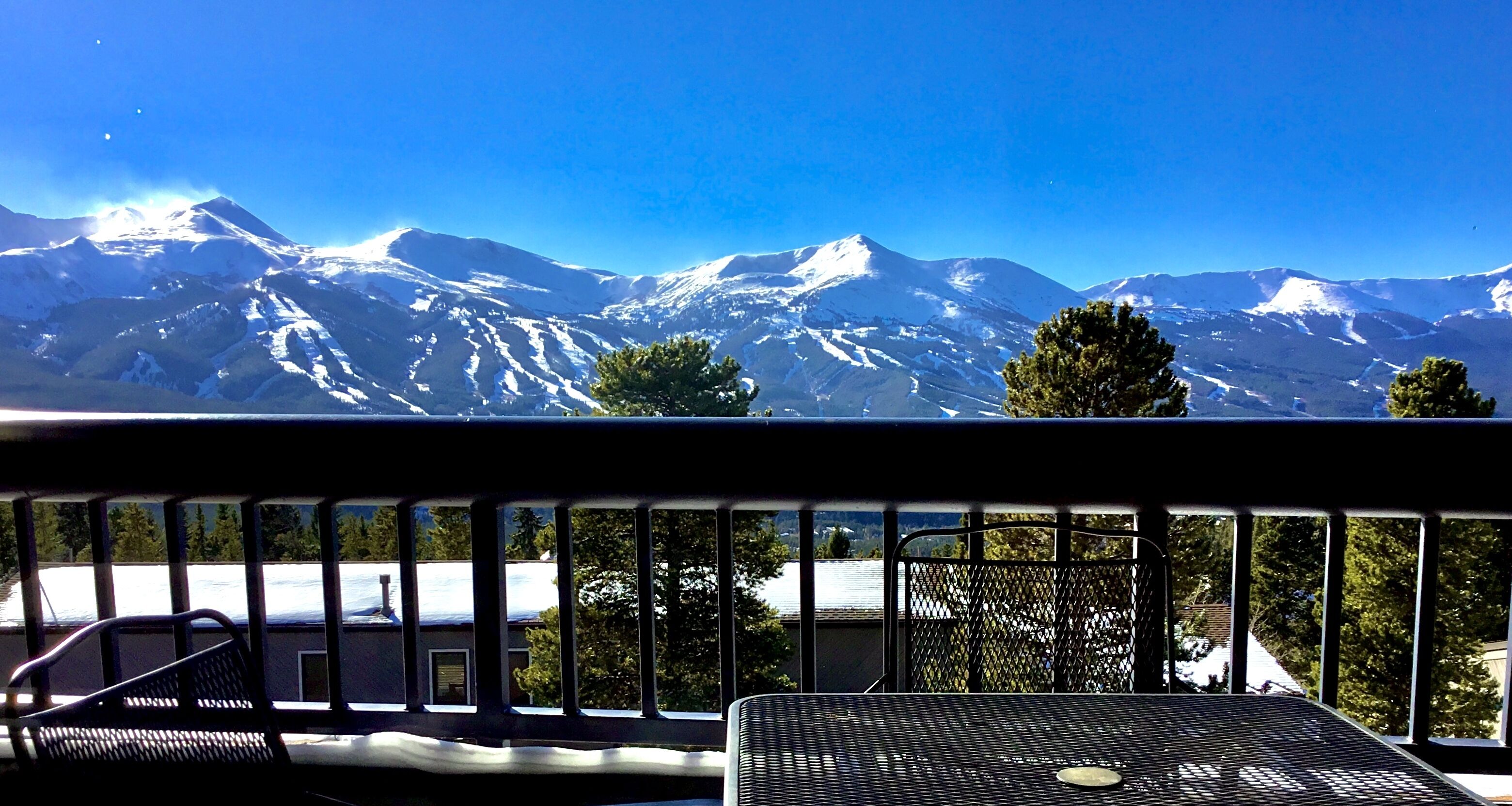 Un balcón con vistas a montañas nevadas, barandilla metálica y una mesa con una silla.