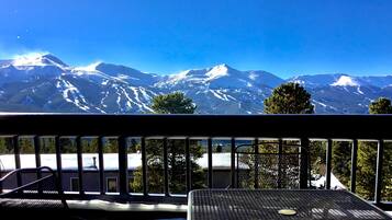Un balcón con vistas a montañas nevadas, barandilla metálica y una mesa con una silla.