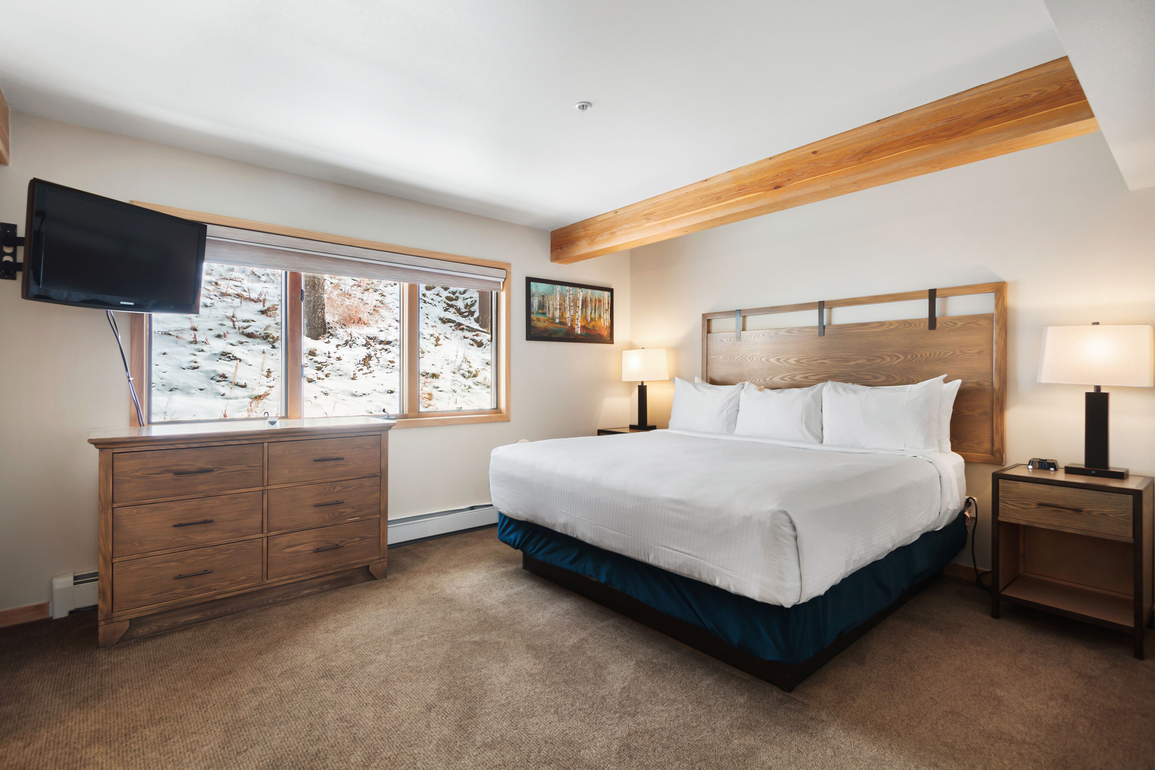 A bedroom with a large bed, a TV mounted on the wall, a window with a view of trees, and a wooden ceiling beam.