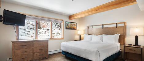 A bedroom with a large bed, a TV mounted on the wall, a window with a view of trees, and a wooden ceiling beam.