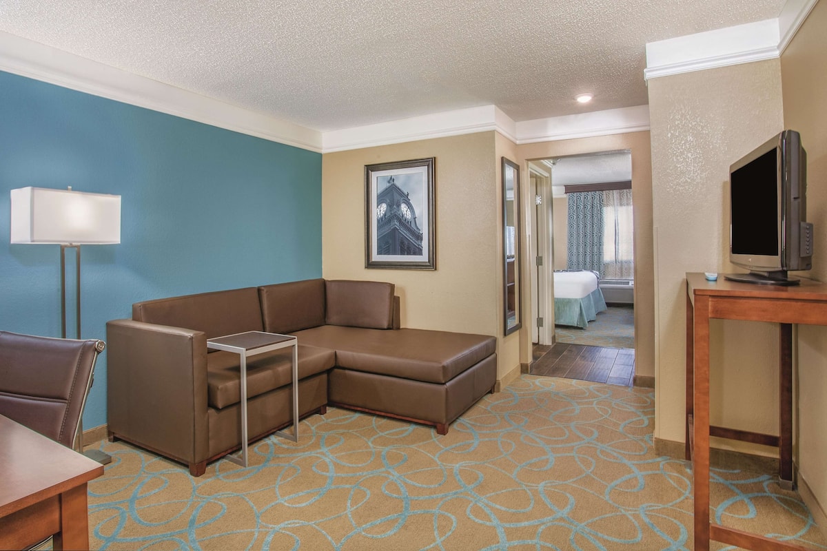 A hotel room with a brown sectional sofa, a small table, a TV on a wooden stand, and a framed picture on the wall.