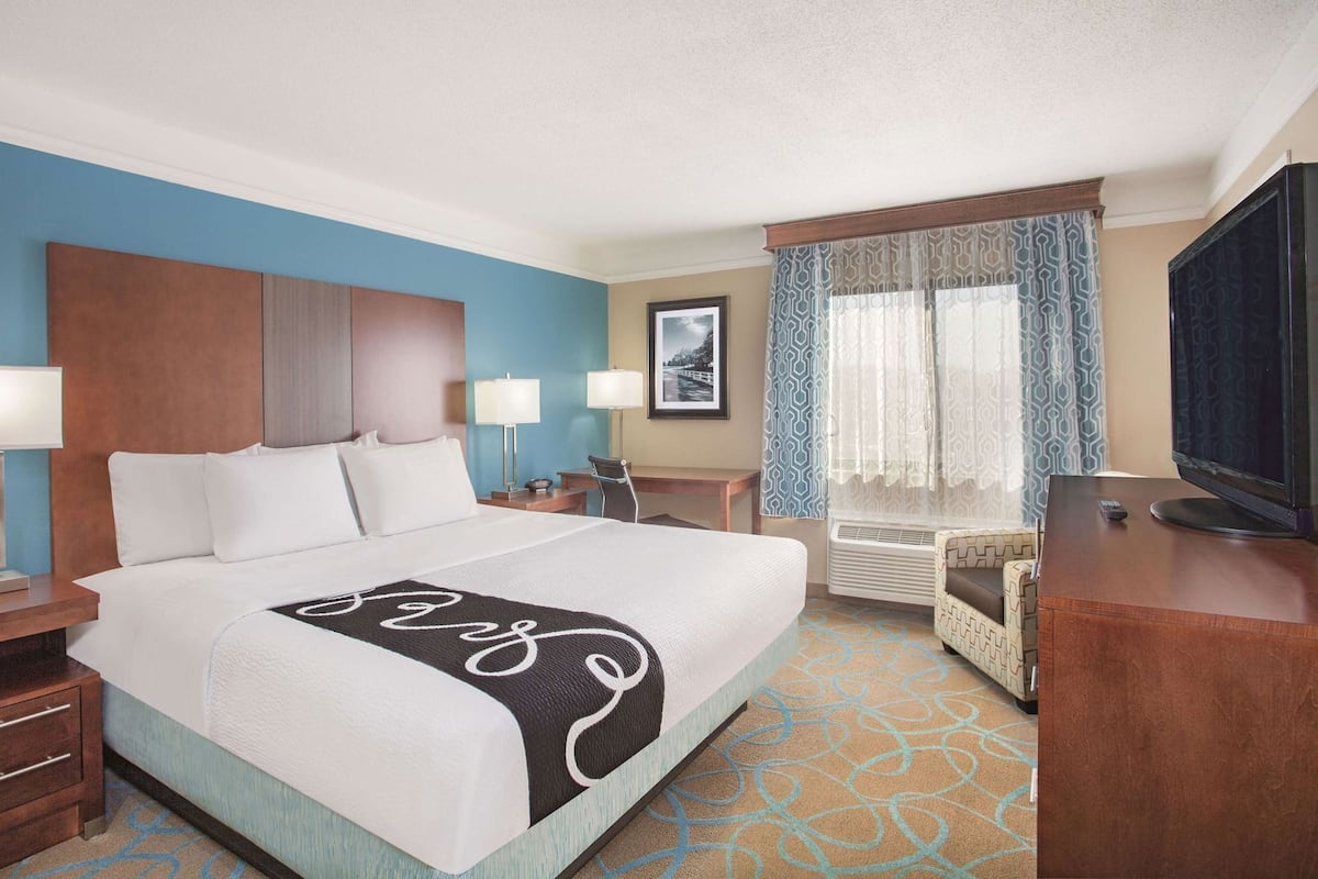 A hotel room with a bed, bedside tables, a desk, a chair, a television, and a window with patterned curtains.