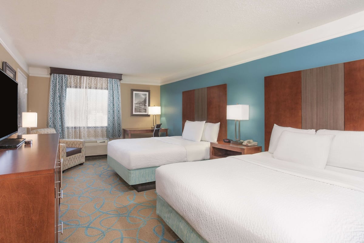 A hotel room with two beds, a desk, a TV, a window with curtains, and a wall-mounted lamp.