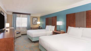 A hotel room with two beds, a desk, a TV, a window with curtains, and a wall-mounted lamp.