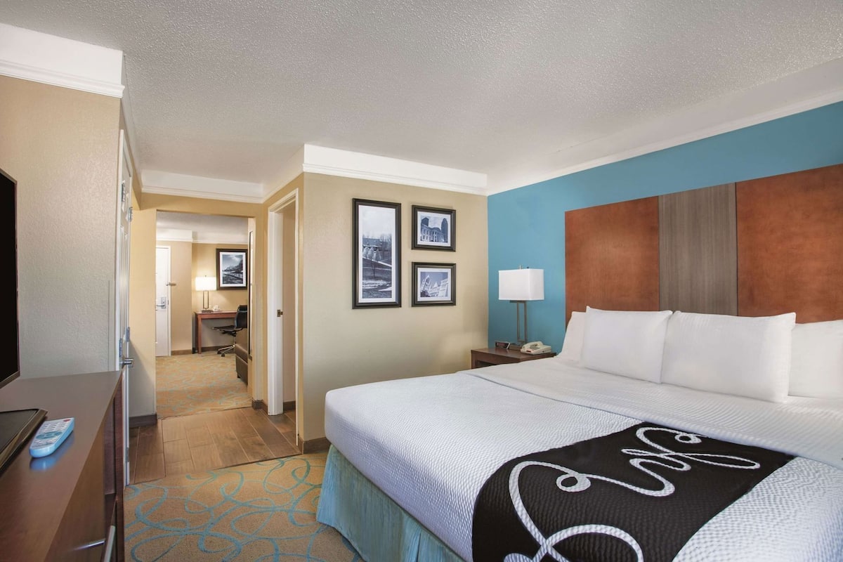 A hotel room with a bed, a desk, a chair, a lamp, and framed pictures on the wall.