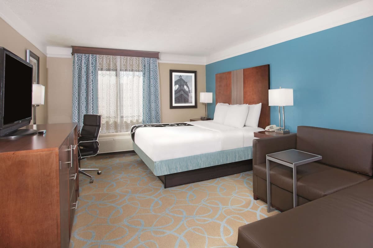 A hotel room with a bed, a desk with a TV, a sofa, a chair, and a window with curtains.