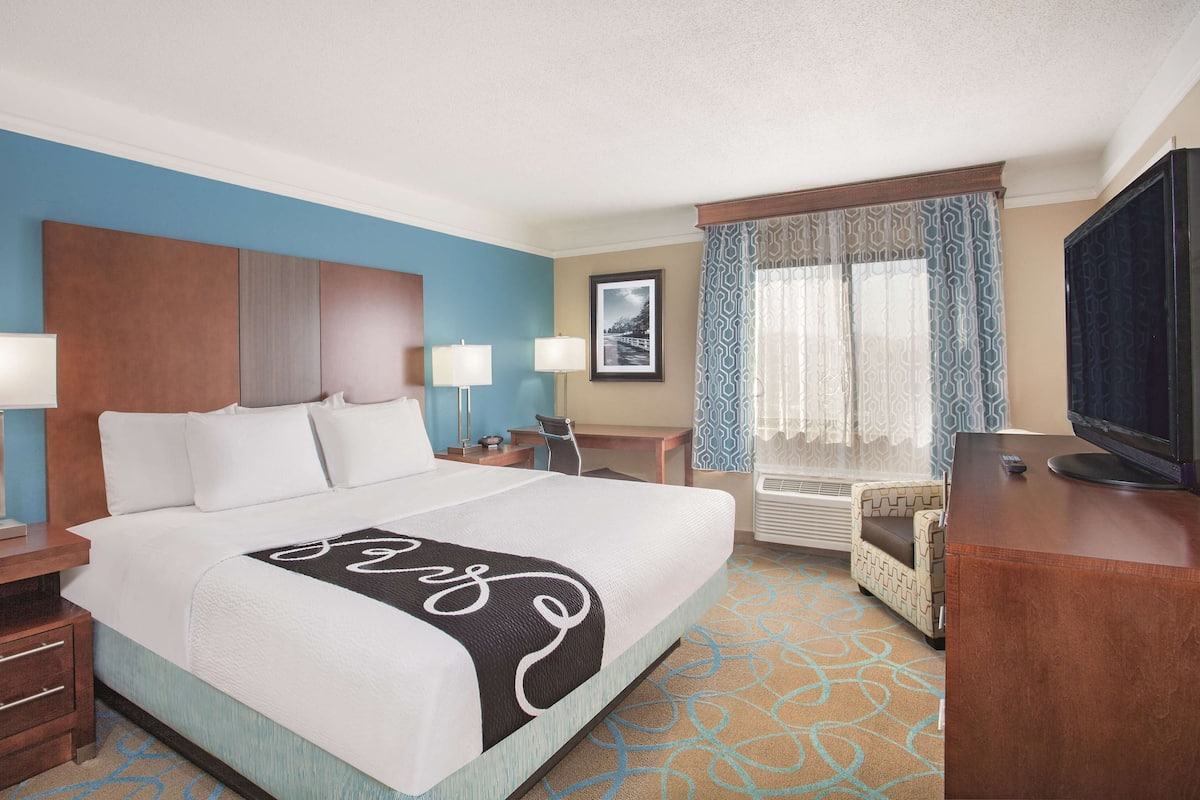 A hotel room with a bed, two bedside tables, a desk, a chair, a television, and a window with patterned curtains.