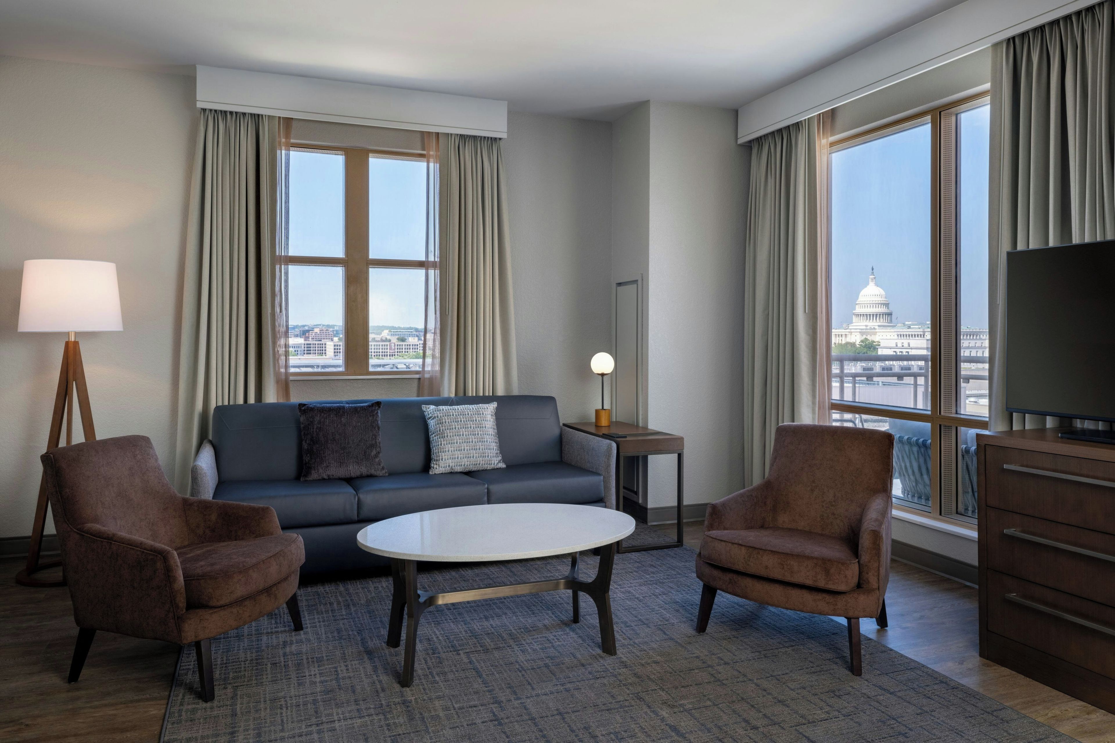 A modern hotel room with a sofa, two armchairs, a coffee table, a lamp, a TV, and large windows offering a city view.