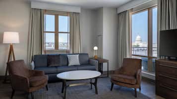 A modern hotel room with a sofa, two armchairs, a coffee table, a lamp, a TV, and large windows offering a city view.