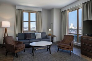 Habitación de hotel moderna con sofá, dos sillones, una mesita de café, lámpara, televisor y ventanas amplias con vista a la ciudad.