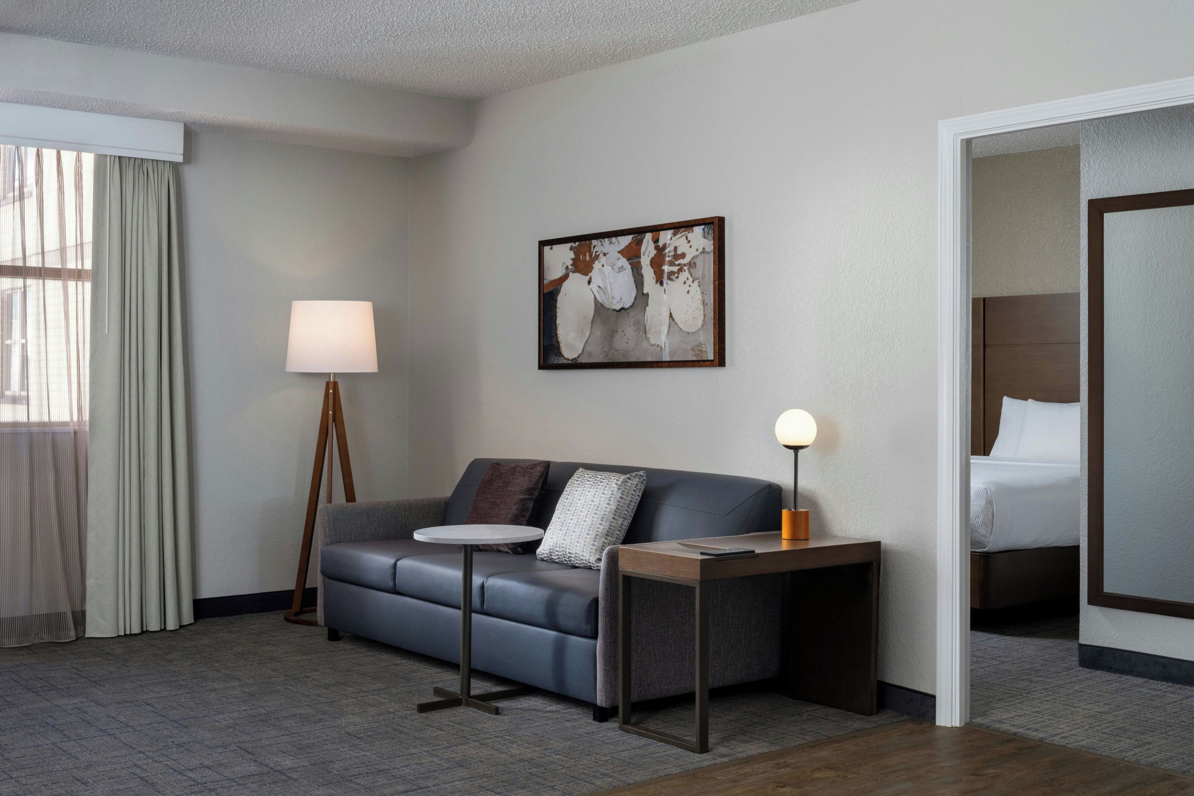Habitación de hotel con sofá, una pequeña mesa redonda, lámpara, un cuadro en la pared y cama en una esquina.