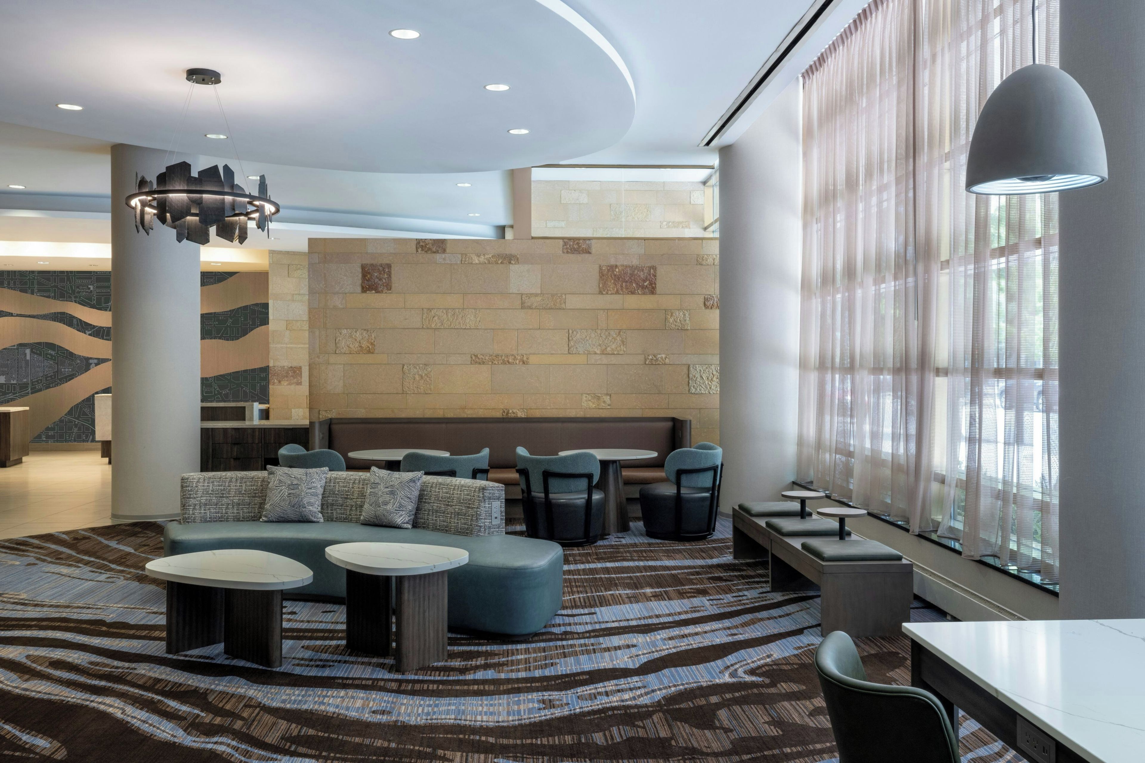 A modern lounge area with seating arrangements, a patterned carpet, and a large window with sheer curtains.