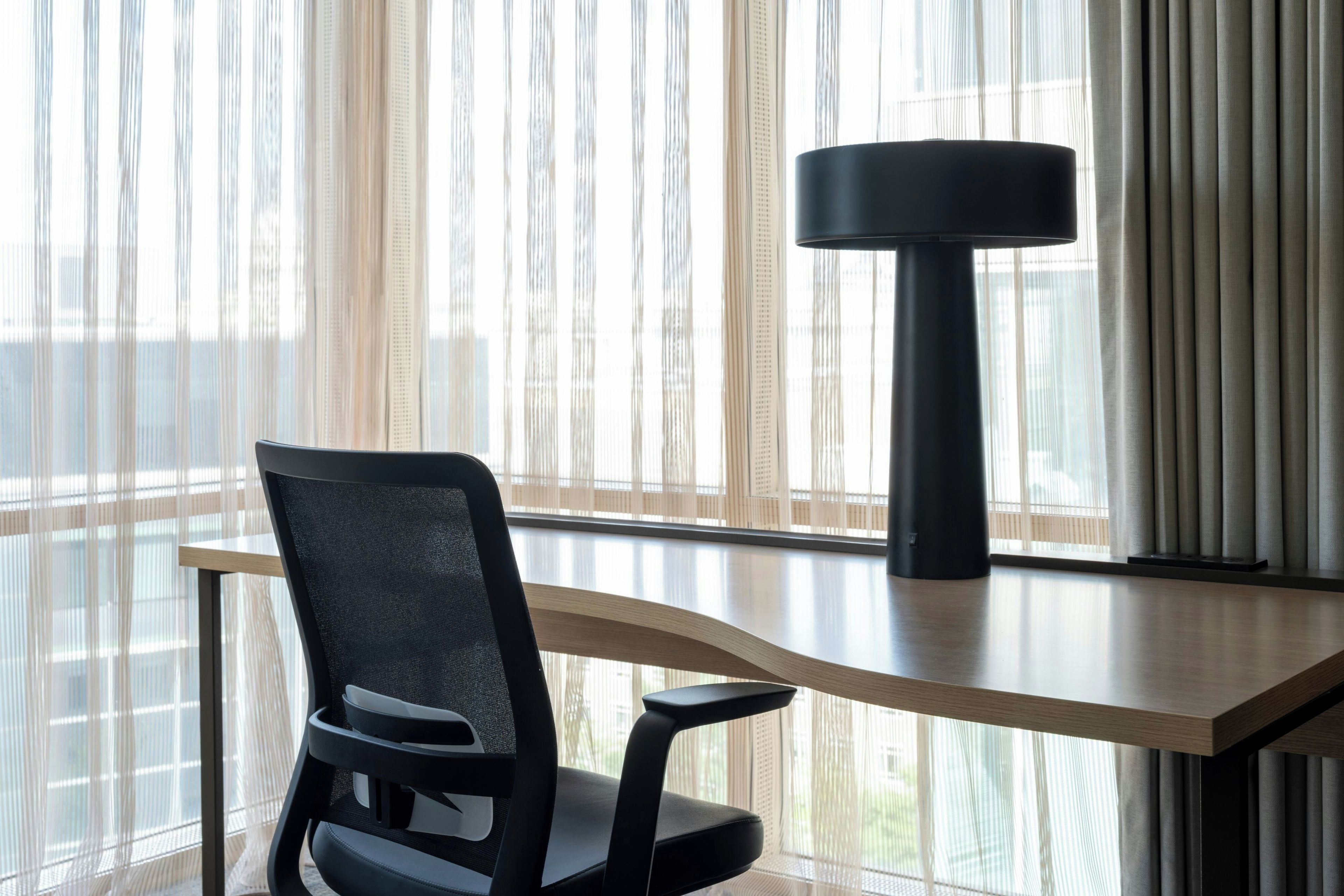 Een moderne bureaustoel met een hoge rugleuning en armleuningen, geplaatst bij een houten bureau waarop een zwart, cilindervormig object ligt, voor een raam met verticale jaloezieën.