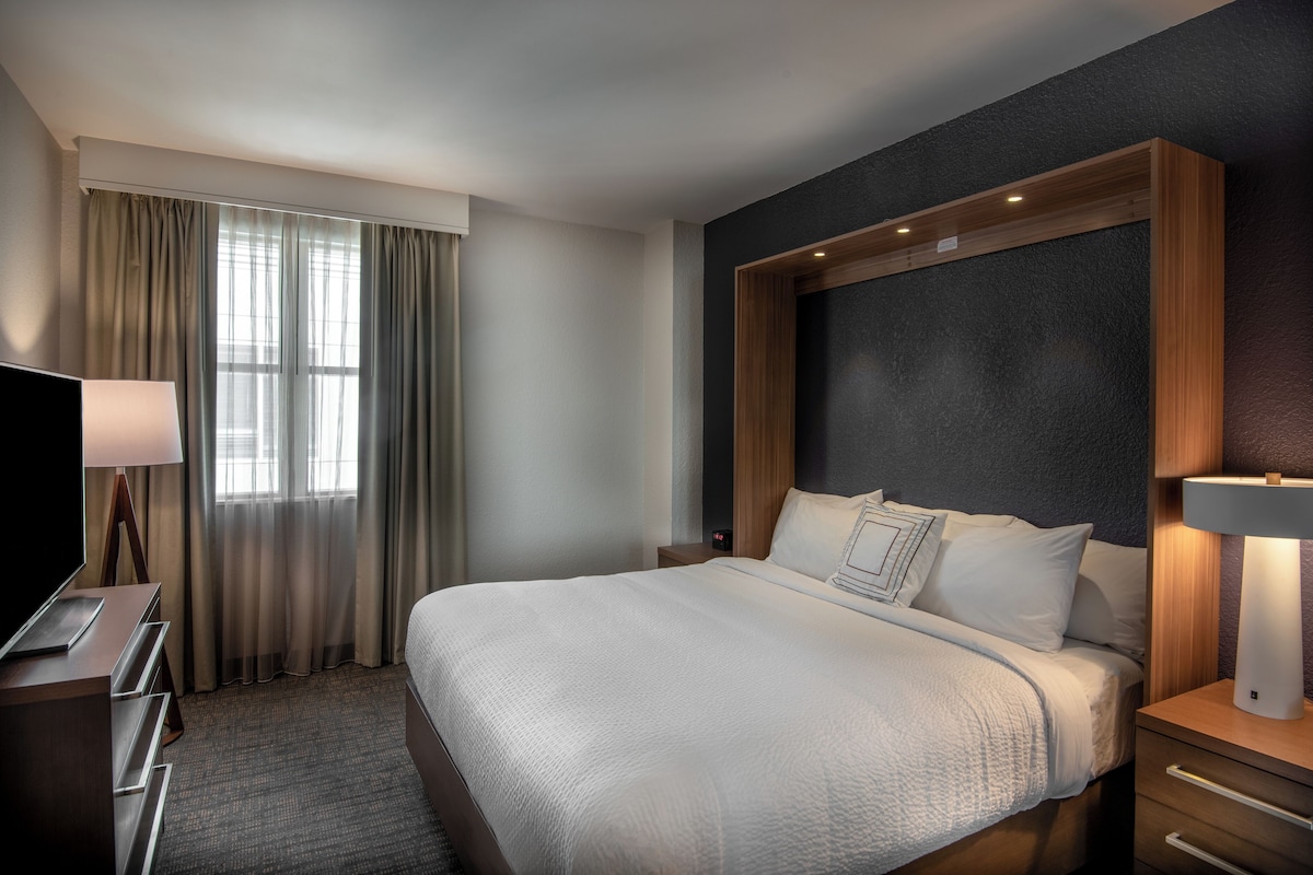 A hotel room with a large bed, a desk with a lamp, a TV, and a window with curtains.