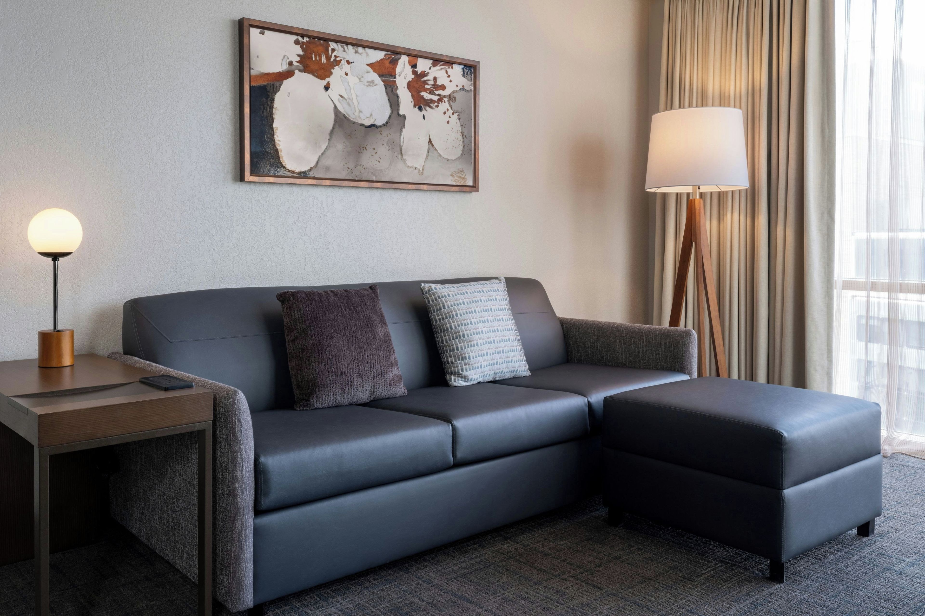 Habitación de hotel moderna con un sofá gris, una mesita de noche con una lámpara y un cuadro abstracto grande en la pared.