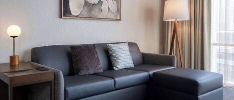 Habitación de hotel moderna con un sofá gris, una mesita de noche con una lámpara y un cuadro abstracto grande en la pared.